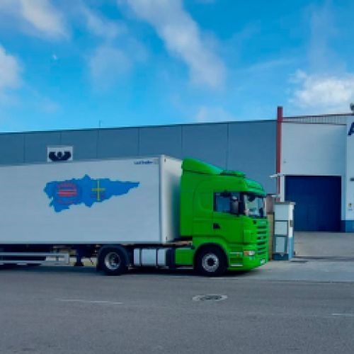 Camión verde con remolque blanco y logotipo aparcado junto a naves industriales